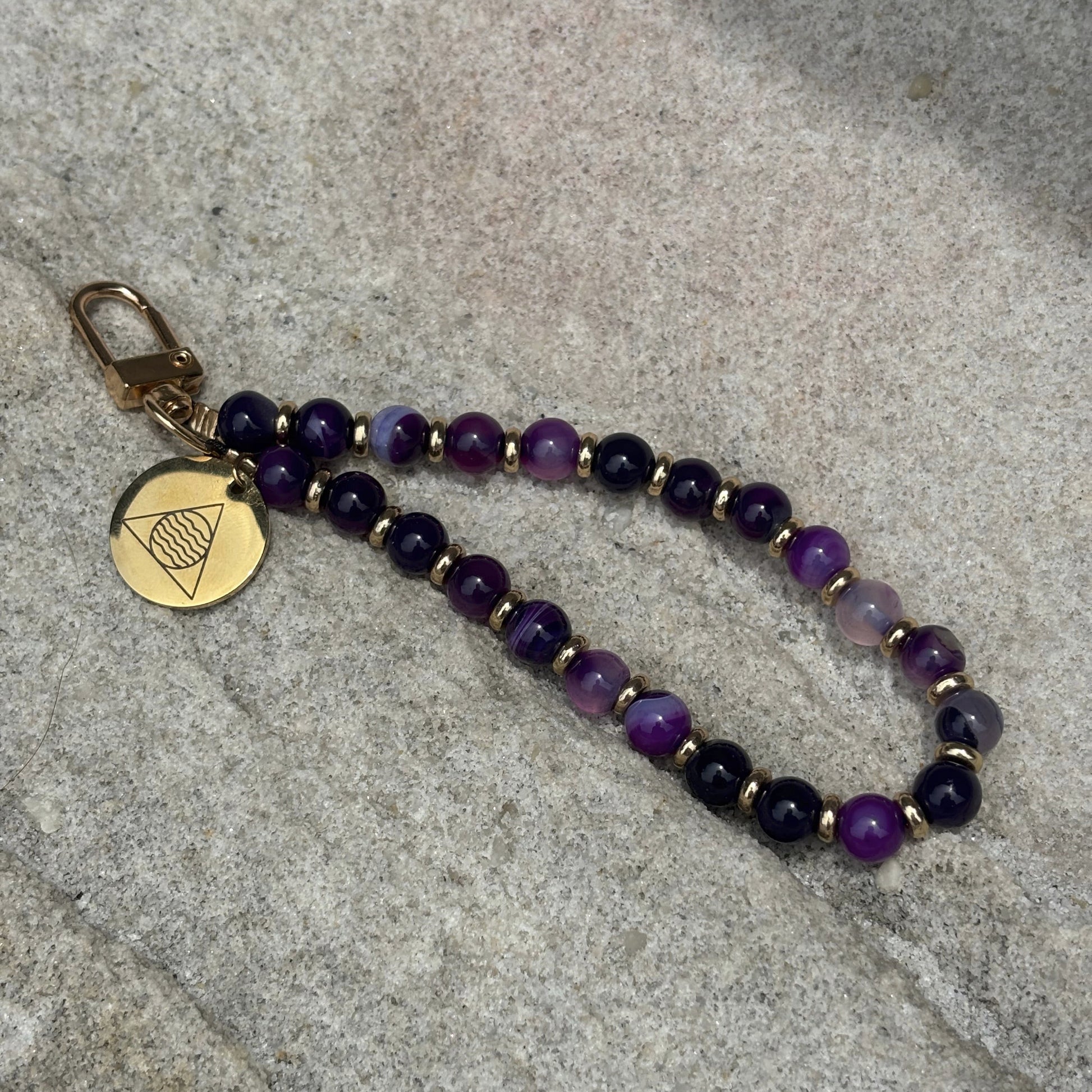 Beaded phone chain with purple and black beads and a gold tag on a stone surface