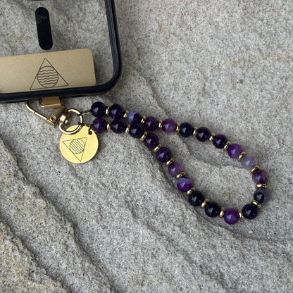 Phone case with a beaded lanyard and gold tag on a stone surface
